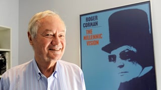 Roger Corman, ein Mann mit grauen Haaren, steht neben einem Filmplakat und lächelt.