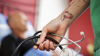 Eine Ärztin mit tätowiertem Arm hält ein Stetoskop in der Hand.