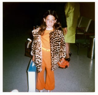 Ein kleines Mädchen im orangefarbenen Overall mit Leopardenmantel schaut in die Kamera.