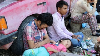 Menschen lehnen erschöpft an ein pinkes Auto an, ein Mädchen schläft auf eines Mannes Schoss.