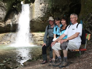 Vier Personen sitzen vor einem kleinen Wasserfall auf einer Holzbank.