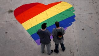 Zwei Personen stehen vor einem regenbogenfarbig auf den Boden gemalten Herzen.