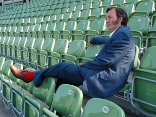 David Pountney sitzt in den grünen Schalenstühle der Seebühne, in blauem Anzug, einen Fuss mit roten Socken über die vordere Sitzreihe gelegt.