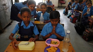 Indisches Klassenzimmer mit zwei Schülerinnen im Vordergrund; eine betet vor dem Essen.
