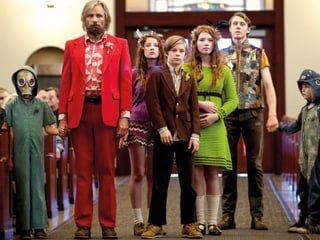 Ein Mann im roten Anzug mit sechs bunt gekleideten Kindern in einer Kirche.