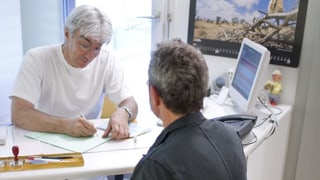Arzt schreibt, Patient sitzt gegenüber. Arzt schreibt, Patient sitzt gegenüber.