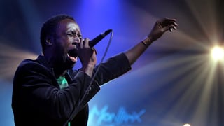 Saul Williams singt auf der Bühne. Er hält eine Hand in die Höhe. Saul Williams singt auf der Bühne. Er hält eine Hand in die Höhe.