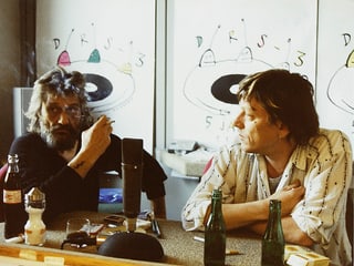 Zwei Männer an einem Tisch, auf dem unter anderem Bierflaschen stehen.