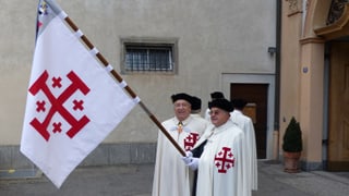 Männer in weissen Roben tragen eine Fahne mit einem aufgestickten Jerusalemkreuz.