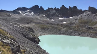 In einer Berglandschaft ist im Vordergrund ein milchig-bläulicher Gletschersee zu sehen. Dahinter eine Gipfelreihe, beim höchsten Gipfel ist ein kleiner Gletscher zu sehen.