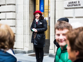 Eine Frau mit rotem Hut steht vor einem Gebäude. Vor ihr sind Menschen.