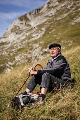 Mann sitzt mit Stock auf der Alp im Gras.