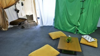 Eine provisorische Unterkunft mit Sitzkisten auf dem Boden und einem Bett in der Ecke.