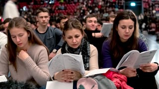 Drei junge Frauen sitzen in einem grossen Raum mit anderen, sie halten Papiere in der Hand. Drei junge Frauen sitzen in einem grossen Raum mit anderen, sie halten Papiere in der Hand.