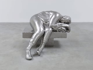 Eine silbrige Skulptur zeigt eine Frau, die auf einer Bank sitzt und ihren Oberkörper zum Schlafen zur Seite gelegt hat. 