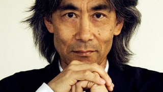 Porträt Kent Nagano, er hat die Hände vor seinem Gesicht übereinandergelegt. Trägt einen schwarzen Kittel.