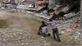 Ein Junge schiebt ein anderes Kind in einem Kinderwagen durch die Strassen der zerstörten Stadt Homs.