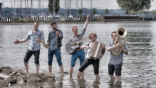 Die fünf jungen Musiker stehen mit kurzen Hosen im Wasser am Ufer eines Sees.