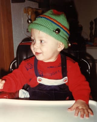 Ein kleine Bub mit grüner Mütze und rotem Pullover sitzt in einem Kindersitz an einem Tisch.