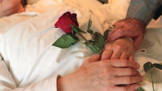 Eine Frau liegt in einem Bett und hälte eine rote Rose auf ihrem Bauch - zusammen mit Männerhänden.