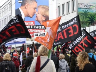 Demonstranten vor einem Schild, auf welchem sich Obama und Merkel küssen.