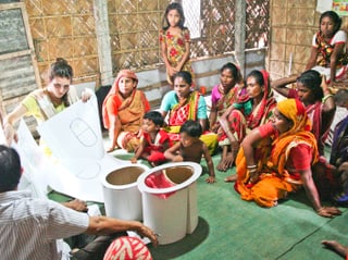 Eine weisse Frau zeigt bunt gekleideten Frauen aus Bangladaseh die Baupläne einer Toilette.