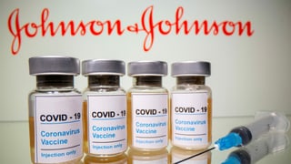 Im Vordergrund Impfdosen von Johnson&Johnson und eine Impfspritze, im Hintergrund das Logo von Johnson&Johnson