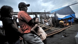 Ein Mann steht mit zwei Kindern vorne, im Hintergrund wird sein kaputtes Haus wieder aufgebaut.