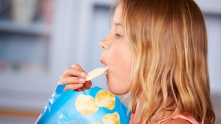 Ein Mädchen mit einer Tüte Pommes-Chips schiebt sich eine Scheibe in den Mund.