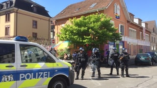 Polizeiauto und Polizisten, dahinter ein Haus mit Baum davor und Sonnenschirm