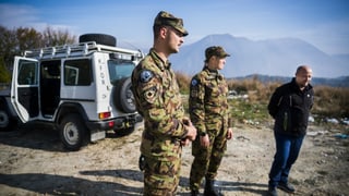 Zwei Swisscoy-Soldaten in Tarntenue stehen neben einem Zivilisten, dahinter ein weisses Fahrzeug, angeschrieben mit KFOR.