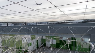 Stacheldraht im Flughafengefängnis von Zürich-Kloten – darüber ist ein Flugzeug am Himmel zu sehen