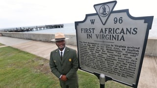 Mann steht vor Tafel. Darauf steht: First Africans in Virginia. Mann steht vor Tafel. Darauf steht: First Africans in Virginia.