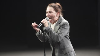 Eine Frau im Anzug spricht mit weit geöffnetem Mund und ernstem Blick in ein Mikrofon. 
