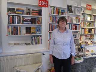 Dorothee Neubauer, kurze Haare, Brille und helle Brille steht vor den Bücherregalen in ihrem Geschäft.