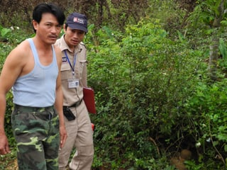 Ein Mann in Unterhemd und einer in beiger Uniform neben einem Gebüsch, in dem eine Hutgrosse Bombe liegt.