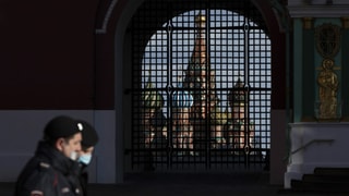 Personen mit Mundschutz gehen an einem vergitterten Tor vorbei. Im Hintergrund ist eine russisch-orthodoxe Kirche zu sehen.