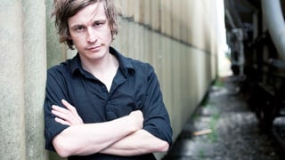 Autor Arno Camenisch lehnt sich im schwarzen Hemd an eine Wand.