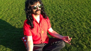 Ein Mann mit langen schwarzem Haar, weit geöffnetem Hemd und Sonnebrille sitzt im Lotussitz auf einer Wiese