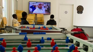 Symbolbild: Asylbewerber sitzen in einer Unterkunft vor einem Fernseher, im Vordergrund ein «Töggeli-Kasten».