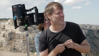 Mann mit kürzeren, braunen Haaren steht neben einem filmtechnischen Gerät. Hinter ihm ist die Stadt Matera zu sehen. Mann mit kürzeren, braunen Haaren steht neben einem filmtechnischen Gerät. Hinter ihm ist die Stadt Matera zu sehen.