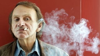 Ein Mann mit schütterem blonden Haar steht rauchend vor einer roten Wand.