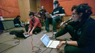 Musiker sitzen am Boden, vor ihnen Laptops, Verstärker, in der Ecke ein Schlagzeuger. Musiker sitzen am Boden, vor ihnen Laptops, Verstärker, in der Ecke ein Schlagzeuger.