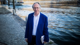 Der britische Schriftsteller Ian McEwan steht mit einem Buch unter dem Arm am Ufer eines Flusses.