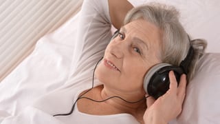 Eine ältere Frau mit Kopfhörern über den Ohren liegt in einem weiss bezogenen Bett.