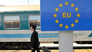 Ein Mann geht an einem Schild mit dem EU-Logo entlang, im Hintergrund ein Zug.