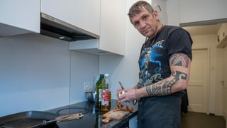 Ein Mann schneidet in der Küche eine Zwiebel und blickt in die Kamera. Am Unterarm ist er tätowiert. Ein Mann schneidet in der Küche eine Zwiebel und blickt in die Kamera. Am Unterarm ist er tätowiert.