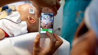Eine Person mit OP-Kleidung macht ein Handyfoto eines Säuglings mit Atemschläuchen in einem Spital.