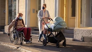 Eine Frau mit Kinderwagen spaziert auf einem Trottoir neben einem Mann im Rollstuhl.