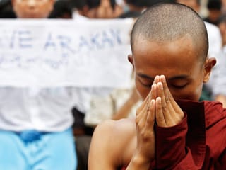 Ein buddhistischer Mönch steht mit gefalteten Händen und gesenktem Blick vor einem Protestplakat.